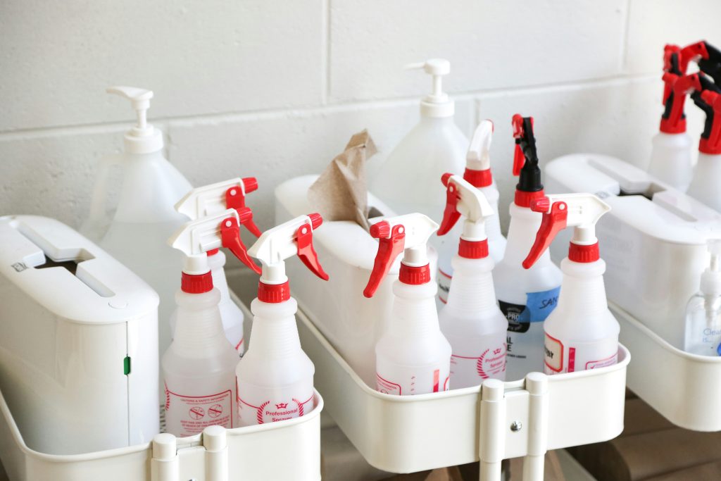 A selection of chemical spray bottles and bulky plastic chemical containers arranged on a shelf, used for cleaning or industrial purposes; labels visible with safety warnings.